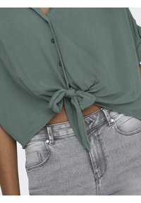 Korte mouwen, top met strik in gedempt groene stof met een gemêleerde afwerking, gecombineerd met zilvergrijze strakke jeans. Knopen zichtbaar op het shirt.