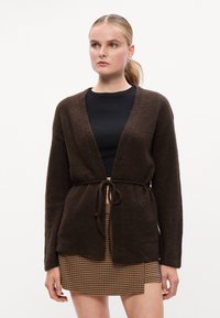 Cárdigan marrón de punto con frente abierto, mangas largas y cinturilla atada. Combinado con una blusa negra ajustada y una falda de patrón de espiga.