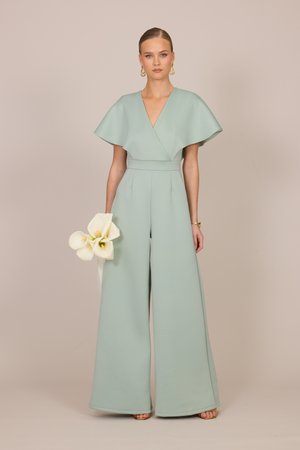 Kvinde iført bredbenet, sart grøn jumpsuit med kappeærmer, der holder hvide calla liljer og står mod en neutral baggrund.