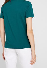 Camiseta de manga corta en color azul verdoso, confeccionada en algodón, con cuello redondo y silueta ajustada, combinada con pantalones blancos. Textura suave, con un detalle mínimo.