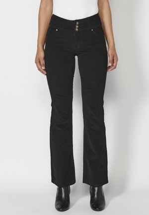 Vrouw draagt zwarte hoge taille corduroy flare broek met drievoudige knoopsluiting en zwarte enkellaarsjes tegen een witte achtergrond.