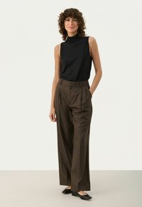 Zwarte mouwloze top gecombineerd met bruine wijde broek met hoge taille en geplooid ontwerp. Model staat op zwarte platte schoenen.