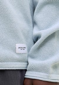 Manche de pull tricoté bleu clair avec poignet replié et étiquette de marque Jack & Jones blanche cousue près de l'ourlet au-dessus d'un pantalon foncé.