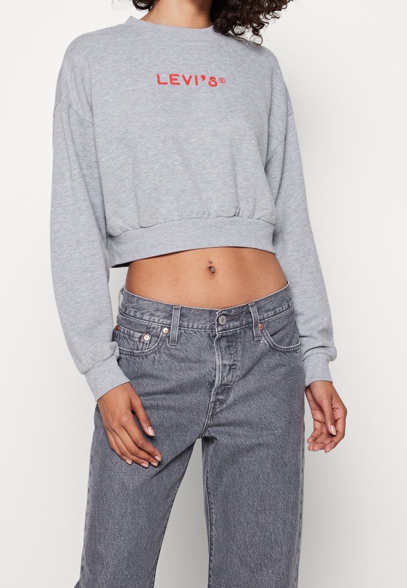 Femme portant un sweat-shirt gris court Levi’s et un jean gris taille haute, montrant le ventre et un piercing au nombril, debout devant un fond blanc.