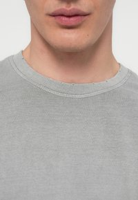Close-up of a person’s lower face and neck wearing a light gray crewneck shirt with slight fraying at the collar.