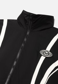 Black jacket with a high collar, featuring white vertical stripes and a round logo patch. The fabric appears smooth and lightweight.