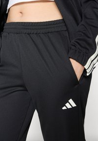 Calças de treino pretas com cintura elástica, apresentando um logótipo branco da Adidas e bolsos laterais. Textura suave com um design desportivo.