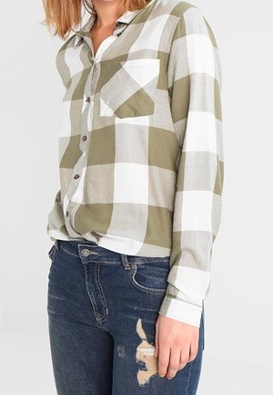 Person wearing a green and white checkered button-up shirt with a front pocket and distressed dark blue jeans.