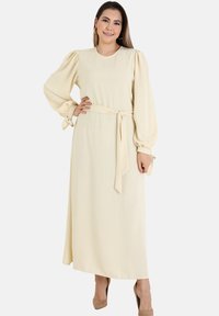 Long, cream-colored dress with puff sleeves and a belted waist. Made from a soft fabric, featuring tie accents at the sleeve hems.