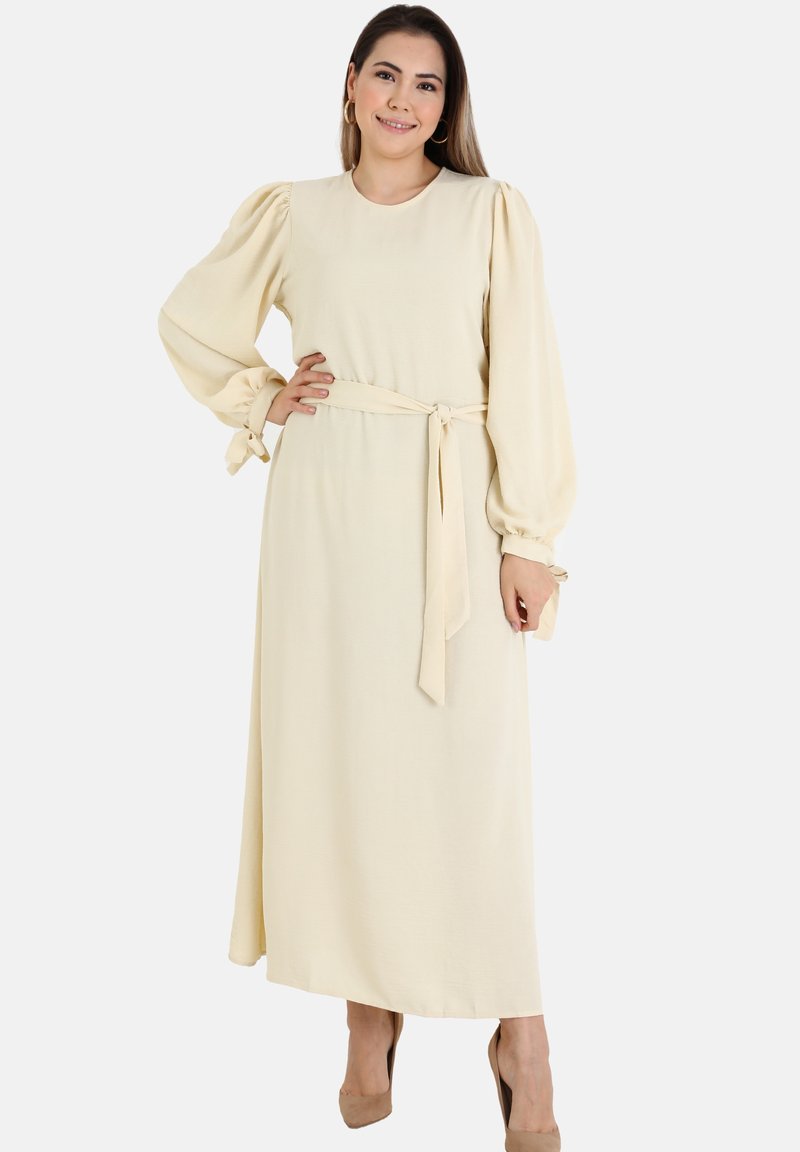 Long, cream-colored dress with puff sleeves and a belted waist. Made from a soft fabric, featuring tie accents at the sleeve hems.
