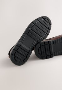 Braune Schuhe mit einer strukturierten schwarzen Gummisohle, die tiefe Profilmuster und eine abgerundete Spitze aufweist. Das Obermaterial zeigt glatte Lederdetails.