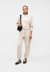 Γυναίκα στέκεται μπροστά σε λευκό φόντο φορώντας μπεζ φούτερ και παντελόνι φόρμας, μαύρη τσάντα χιαστί και μαύρα-άσπρα αθλητικά παπούτσια.