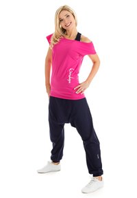 Žiarivo ružový off-shoulder top s bielym logom, skombinovaný s voľnými tmavomodrými teplákmi a bielymi atletickými topánkami.