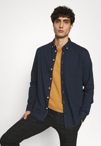 Marineblaue Hemdjacke mit Holzknöpfen, über einem senffarbenen T-Shirt getragen. Schwarze karierte Hose mit dezentem Schachbrettmuster.