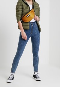 Persona que lleva una chaqueta acolchada verde, camiseta blanca, vaqueros ajustados de tiro alto azules con cremallera frontal, zapatillas en blanco y negro, y un bolso bandolera naranja.