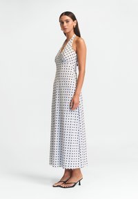 Vit halterneck-klänning med svarta prickar, golvlång, med figurnära bodice och flödande kjol. Har en slät textur och en enkel design.