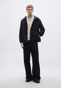 Námornícka vetrovka so zipsom vpredu, elastickými manžetami a kapucňou, vrstvená cez svetlo béžovú košeľu a skombinovaná so širokými tmavými rifľovými nohavicami.