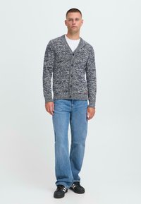 Man som står klädd i en vit t-shirt, en mörkblå och vit melerad cardigan med knappar, blå jeans och svarta sneakers med vita ränder.