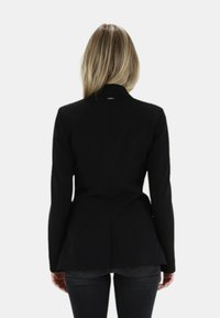 Blazer nero su misura con vita aderente, maniche lunghe, spacco singolo sul retro e texture liscia. Abbinato a jeans scuri.