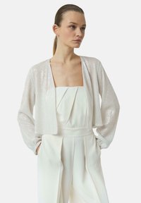 Kurze, taillierte Jacke mit schimmernder silberner Textur, ausgestattet mit langen Ärmeln und offenem Frontdesign, kombiniert mit einem weißen Overall.