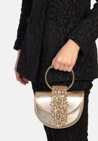 Borsa dorata dal design strutturato, con un manico circolare, decorata con pietre preziose trasparenti lungo la parte frontale. Abito nero dalla texture visibile.