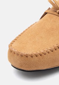 Nærbillede af lysebrun ruskindsmoccasin med syninger langs den afrundede tå og den øverste kant, med en sort sål synlig nedenunder.