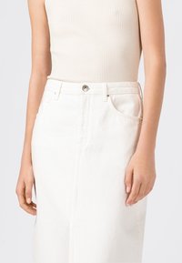 Bílá denimová sukně s předními kapsami, poutky na opasek a kovovým zapínáním. Tkanina má mírně texturovaný povrch.