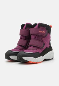 Lila und schwarze Kinderstiefel mit Klettverschluss, strukturiertem Obermaterial und einer stabilen weißen Sohle mit orangenen Akzenten. Geeignet für aktive Nutzung.