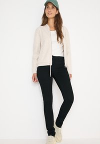 Chaqueta de cremallera beige clara sobre una camiseta blanca, combinada con vaqueros skinny negros y zapatillas beige. La gorra es de color verde claro.