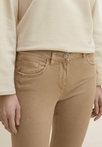 Jeans elásticos beige con un diseño clásico de cinco bolsillos, que presentan un cierre frontal con botón y detalles de costura sutiles. Textura suave y lisa.