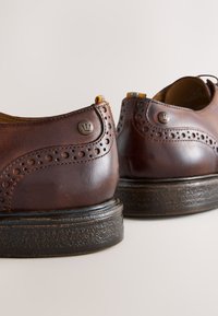 Brune lædersko med brogue-detaljer, rund form, teksturerede såler og en lille metalaccent nær hælen.