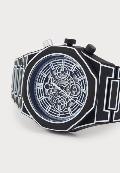 Orologio ottagonale nero con movimento meccanico trasparente. Accenti bianchi mettono in evidenza il design, caratterizzato da linee audaci e due pulsanti.