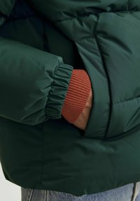 Mørkegrønne puffjakke med ribbet, rustfarvet manchet. Har en sidelomme, glat tekstur og samlet ærmedetalje.