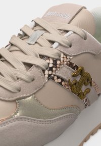 Μπεζ και μεταλλικό αθλητικό παπούτσι με λεπτομέρειες σε σχέδιο φιδιού, μπεζ κορδόνια και χρυσό έμβλημα φίδι στο πλάι, λογότυπο "Just Cavalli" στη γλώσσα.