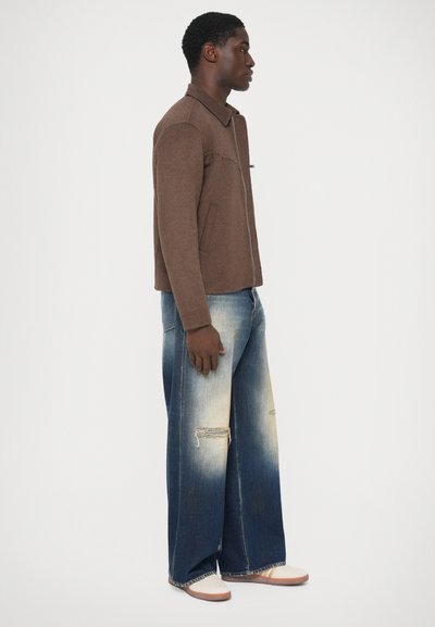 Uomo che indossa una giacca marrone con zip, jeans a gamba larga di un blu sbiadito con strappi, e sneakers bianche, in piedi di profilo su uno sfondo bianco.