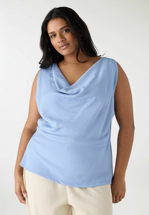 Donna con lunghi capelli scuri che indossa una maglia senza maniche azzurro chiaro con collo a scialle e pantaloni color crema, in piedi davanti a uno sfondo chiaro e uniforme.