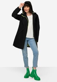 Imperméable noir avec une capuche, accents de fermeture éclair vert fluo et poches avant, associé à un jean bleu clair et des bottines vertes brillantes.