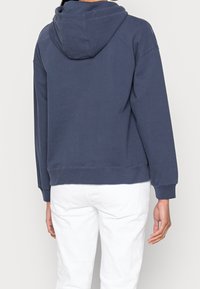 Felpa blu navy con un grande cappuccio, polsini e orlo a coste, realizzata in tessuto di cotone. Pantaloni bianchi parzialmente visibili nella parte inferiore.
