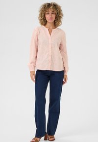 Light pink, long-sleeve blouse with floral embroidery and button front. Paired with dark blue jeans and brown heeled sandals. Smooth texture.