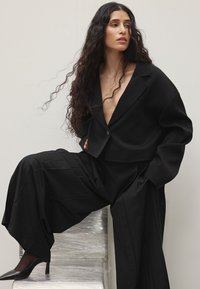 Must musterniga mustri asukate musti mustri mustri vöödiga, millel on sügav V-kaelus ja üks nupuvajutus, koos laia säärega mustade pükste ja punktiga mustade kontsadega.