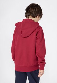 Ein Kind trägt einen roten Kapuzenpullover, schaut mit dem Rücken zur Kamera und hat eine dunkelblaue Hose an, während es vor einem schlichten weißen Hintergrund steht.