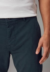 Pantalones de algodón ajustados en un verde oscuro, con una textura suave. Cuentan con un cierre de botón, bolsillos laterales y una cinturilla ajustada.