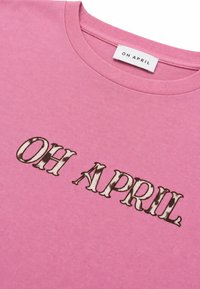 Ροζ μπλουζάκι με στρογγυλή λαιμόκοψη και "OH APRIL" τυπωμένο με μεγάλα, στιλιζαρισμένα γράμματα στο στήθος και ετικέτα μάρκας στο λαιμό.