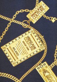 Maillons de chaîne en or avec des étiquettes rectangulaires Versace Jeans Couture sur un tissu foncé, comportant du texte et le nom de la ville de Milan.