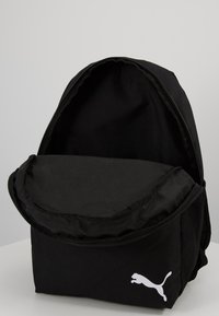 Zaino nero con parte superiore arrotondata, chiusura con zip e tasca in rete frontale. Presenta un evidente logo Puma bianco. Realizzato in tessuto resistente.