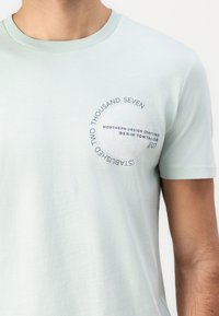 Maglietta corta verde menta con maniche corte, caratterizzata da una grafica circolare con testo nella zona sinistra del petto. Tessuto morbido e liscio.