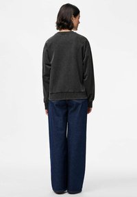 Asmuo su trumpais tamsiais plaukais, atsukęs nugarą, vilkintis išblukusį juodą megztinį ir laisvas tamsiai mėlynas džinsus, priešais paprastą baltą fono.