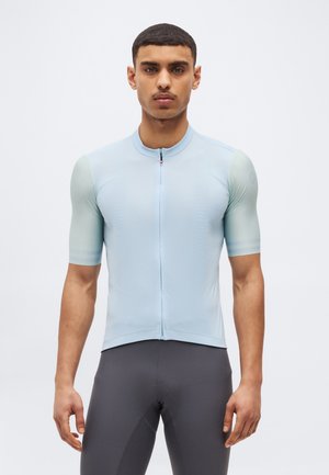 Άνδρας μοντέλο με κοντά μαλλιά, φορώντας ανοιχτό μπλε ποδηλατική φανέλα με κοντά μανίκια και σκούρο γκρι επενδεδυμένο ποδηλατικό σορτς, στέκεται μπροστά σε μονόχρωμο φόντο.