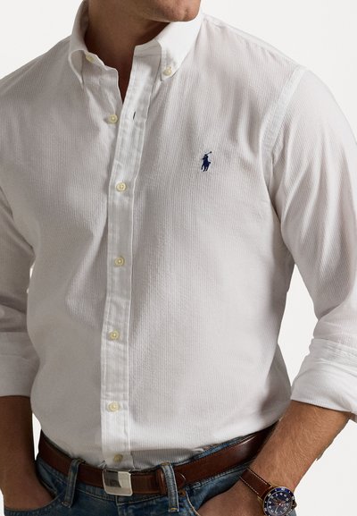 Camisola branca de botões com tecido texturizado, botões amarelos e um logótipo azul no peito. Combinada com jeans de ganga e um cinto de couro castanho.