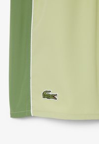 Tissu vert clair et vert foncé avec un liseré blanc et un petit logo de crocodile vert brodé près de l'ourlet inférieur.